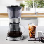 A slim Cold Brew coffee maker with a glass of iced coffee on a countertop.