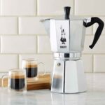 A silver aluminum Moka Express coffee maker on a countertop with two espresso cups.