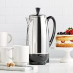 A silver Percolated Coffee maker on a countertop with coffee mugs and a cake.