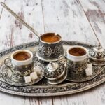 A set of traditional Eastern Black Coffee cups and saucers on a silver tray. A copper coffee pot, is filled with coffee and placed in the center.